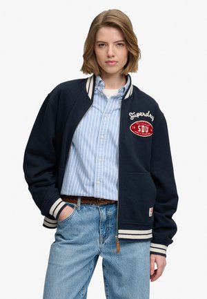 Ung person iført en navy varsity-jakke med hvide stribede manchetter, en blå stribet skjorte med knapper og lyseblå jeans med flettet bælte.