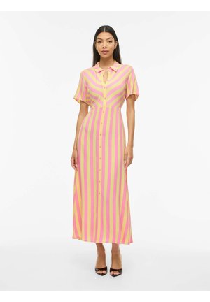 Femme debout portant une longue robe boutonnée à rayures verticales roses et jaunes et manches courtes, associée à des talons ouverts noirs.