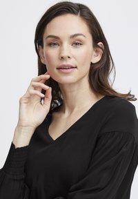 Melna blūze ar V veida izgriezumu un garām pūkām. Tekstūrēta auduma ar smockinga detaļām pie piedurknēm. Vienkāršs, elegants dizains.