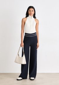 Λευκή αμάνικη μπλούζα με ψηλό γιακά και φερμουάρ, συνδυασμένη με ναυτικές παντελόνες με φαρδιά πόδια που διαθέτουν λευκές ρίγες στη μέση. Κρατάει μια μπεζ τσάντα.