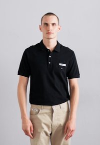 Juodas polo marškinėliai su apykakle, turintys baltą Moschino logotipą kairėje krūtinės pusėje. Audinys atrodo lygus ir lengvas.