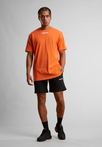 Oransje bomull t-skjorte med "STARTER" logo, sammen med svarte shorts med logo aksenter. Modellen har på seg svarte sneakers og sokker.