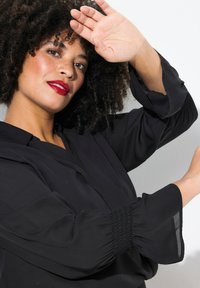 Schwarze Bluse mit langen, glockenförmigen Ärmeln und gesmockten Bündchen. Weicher, glatter Stoff mit lockerer Passform und tiefem V-Ausschnitt.