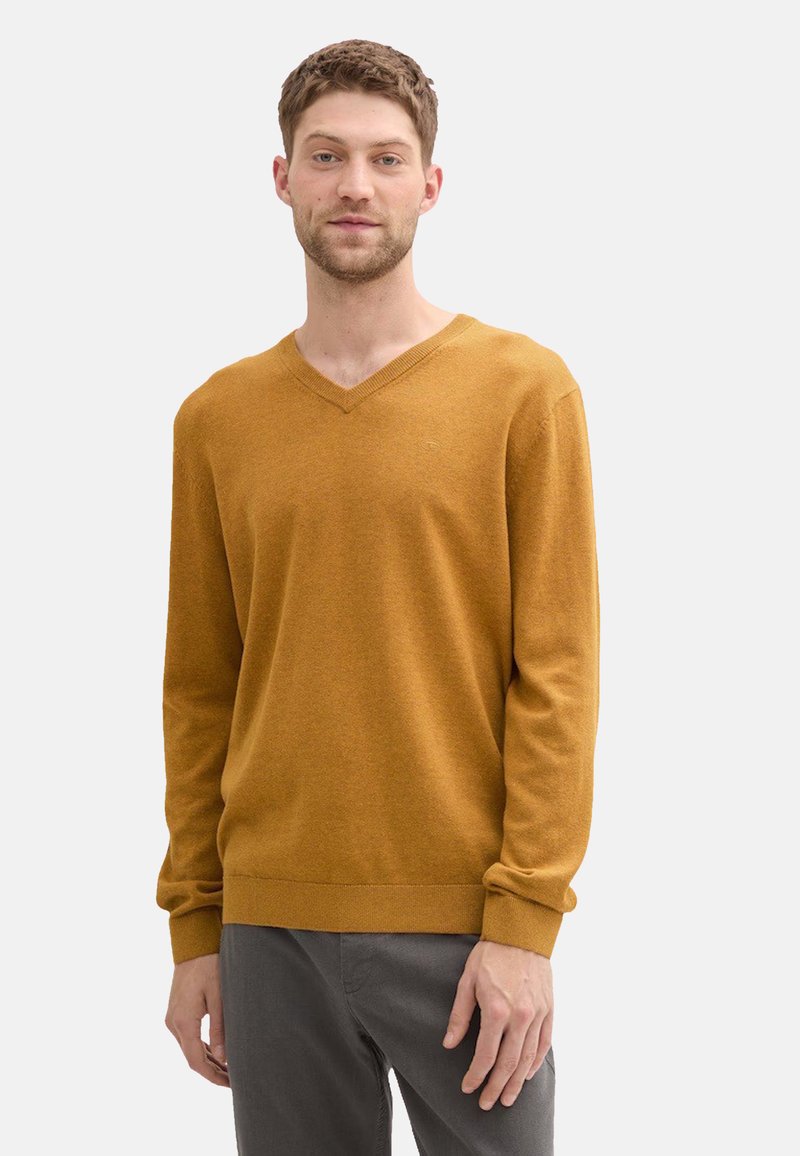 Suéter amarillo mostaza de cuello en v, en una tela suave y con mangas largas. El diseño sencillo presenta un dobladillo y puños acanalados, combinado con pantalones grises.