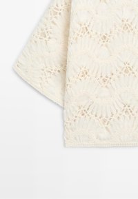 Crèmekleurige stof met gedetailleerde bloemrijke kanten patronen en geschulpte randen op een gevouwen textielstuk.