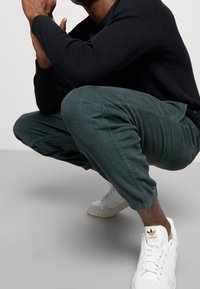 Tmavě zelené manšestrové kalhoty s žebrovanou texturou, doplněné bílými teniskami Adidas se zlatými akcenty. Postava je v podřepu a má na sobě černý pletený svetr.