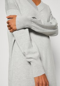 Personne portant une robe-pull en tricot gris clair à manches longues avec des poignets côtelés et un col en V, croisant un bras sur l'autre.