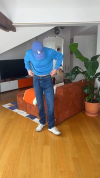 Person iført blå kasket og sweatshirt, blå jeans og hvide sneakers, justerer bukser i stuen med orange sofa og stor potteplante.