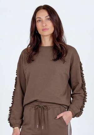 Femme aux longs cheveux bruns portant un sweat-shirt à manches volantées marron et un pantalon à cordon assorti, debout devant un fond uni.