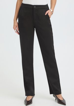 Femme portant un pantalon noir droit, un haut blanc et des talons à bout pointu foncés, avec une main dans la poche et des bijoux visibles.