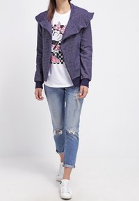 Chaqueta de forro polar morada con un cuello rizado, llevada sobre una camiseta blanca con un personaje de dibujos animados, combinada con jeans azules desgastados y zapatillas blancas.