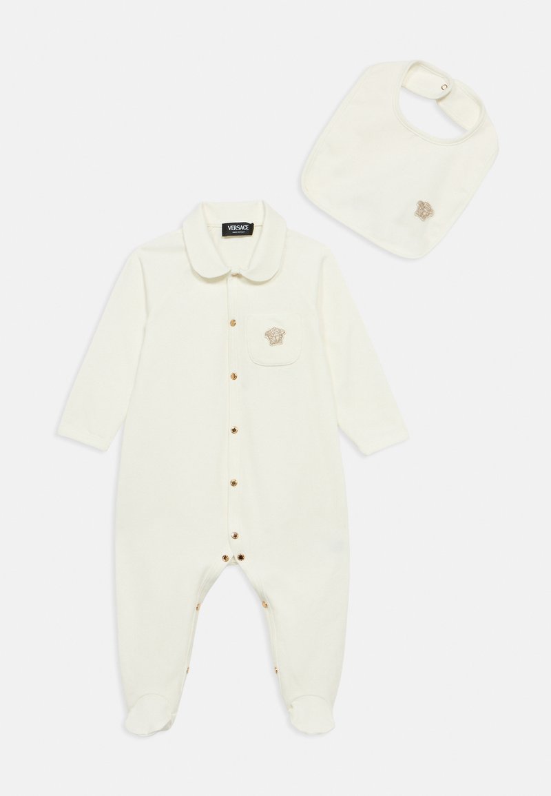 Krém színű babaruházat hosszú ujjakkal, gallérral, elöl gombokkal és zsebbel, amely arany hímzett logót visel; hozzáillő előkével.
