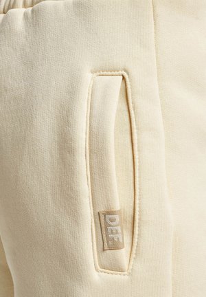 Die crèmefarbenen Jogginghosen verfügen über eine Seitentasche mit einem Markenetikett. Der Stoff hat eine weiche, strukturierte Oberfläche und ein leicht dehnbares Design.