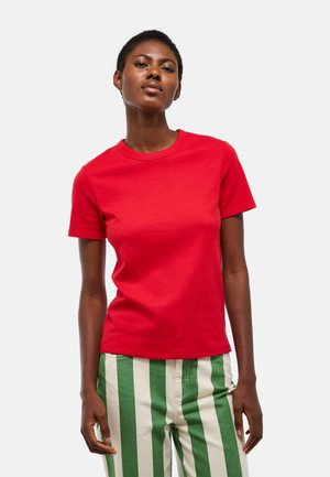 Femme portant un t-shirt rouge uni à manches courtes et un pantalon à rayures verticales vertes et blanches, debout devant un fond blanc.