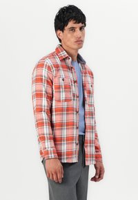 Camicia a quadri rossi e bianchi con dettagli neri, caratterizzata da un design con bottoni, due tasche frontali e una texture morbida, indossata sopra una maglietta blu.
