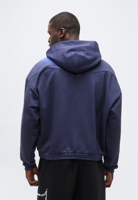 Άνδρας με τη πλάτη, φοράει μπλε σκούρο φούτερ με κουκούλα και μαύρο παντελόνι, σε απλό ανοιχτό γκρι φόντο.