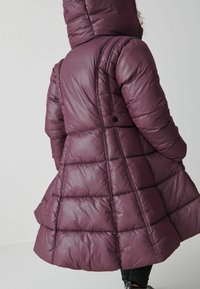 Lång, vadderad vinterjacka i djup burgundy. Har huva, quiltad design och cinchad midja, sträcker sig till mitten av låret med en utställd botten.