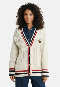 Frau mit lockigem Haar, die eine weiße Strickjacke mit roten, schwarzen und weißen Streifen über einem blauen Hemd und Jeans trägt.