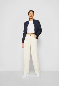 Veste bomber matelassée navy avec des poignets côtelés, associée à un pantalon large crème à coupe ample et des baskets blanches, sur un fond neutre.