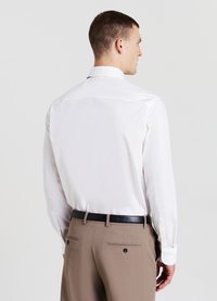 Homme de dos portant une chemise blanche rentrée dans un pantalon beige avec une ceinture noire, sur un fond clair uni.