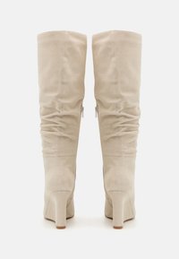 Bottes beiges au-dessus du genou en daim texturé, avec un talon bloc, des détails froncés et des fermetures éclairs latérales.