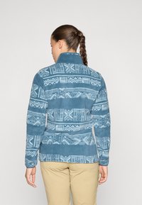 Fleecejakke i teal med geometriske mønstre i lyseblå, høj krave og en tætsiddende pasform. Glat tekstur med lange ærmer.