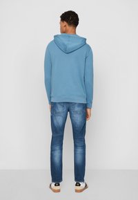 Lichtblauwe hoodie met een trekkoord in de capuchon, gecombineerd met blauwe jeans. De hoodie heeft een gladde textuur; de jeans hebben een lichte vervaging en een klassieke pasvorm.