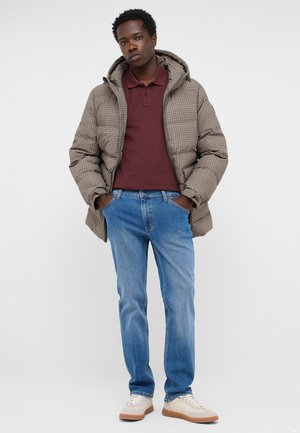 Young man stands with hands in pockets wearing a checked puffer jacket, maroon polo shirt, blue jeans, and beige sneakers on white background.