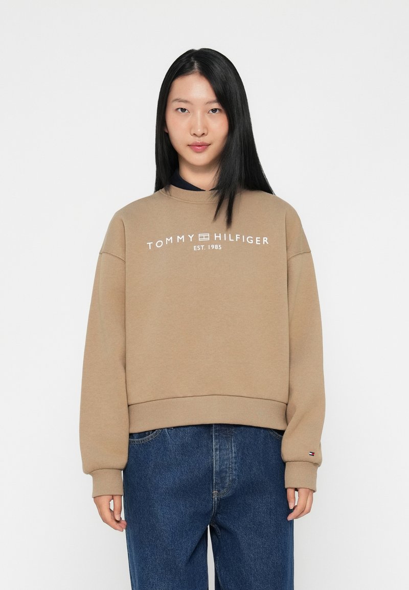 Beige bomullssweatshirt med ribbestrikket rund hals og mansjetter. Hvit logo tekst "TOMMY HILFIGER EST. 1985" sentrert på brystet. Avslappet passform.