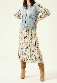 Jeansväst med framfickor lager på en färgglad mönstrad midi-klänning. Knähöga beige stövlar kompletterar outfiten.