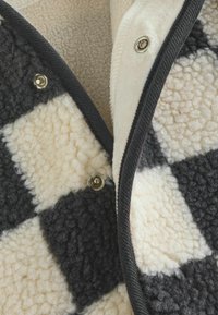 Fleece-takki musta ja kerma värviga ruuduline muster, pehme ja tekstuuriga pind, press-nupud ja kontrastsed tumedad ääred.