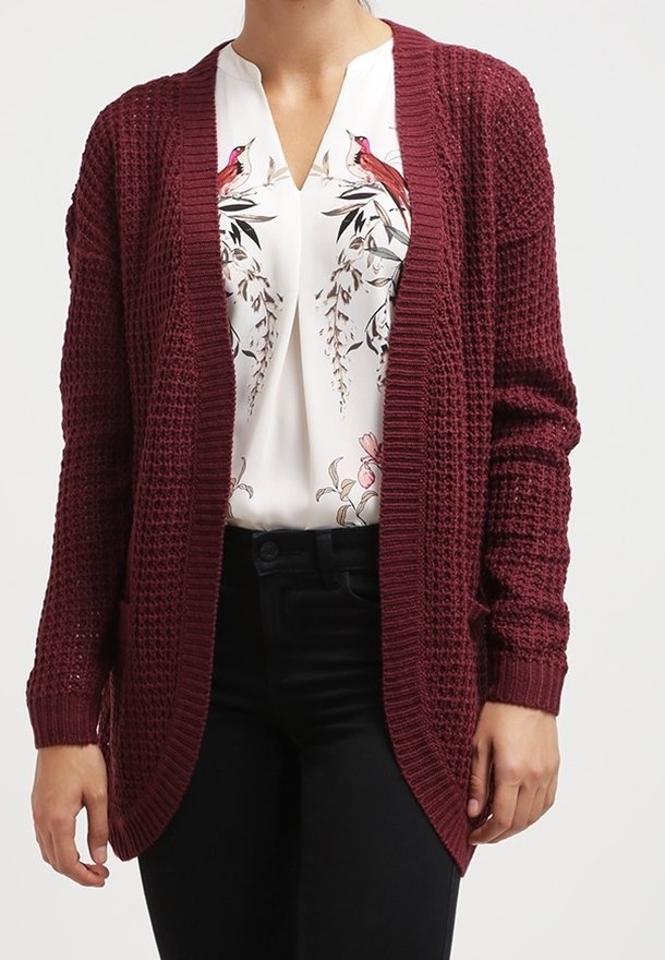 Femme portant un cardigan bordeaux texturé sur une blouse blanche ornée d'imprimés d'oiseaux roses et de motifs floraux, associée à un pantalon noir.
