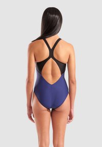 Vrouw met de rug naar de kijker, gekleed in een twee-kleurige blauwe en zwarte badpak met een racerback en een uitsparing op de rug, tegen een effen grijze achtergrond.