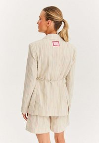 Béžový pruhovaný blazer s odpovídajícími šortkami, které mají zavazovací pas, barevné akcenty a čtvercovou růžovou nášivku na horní části zad.