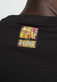 Sort t-shirt med et farverigt, kunstnerisk mærke med "PUMA"-logoet på bagsiden. Stoffet er ribbet, hvilket forbedrer teksturen.