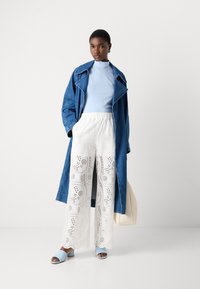 Cappotto trench in denim, top azzurro con colletto alto, pantaloni bianchi a gamba larga con occhiello e sandali blu con zeppa, abbinati a una borsa a spalla crema.