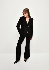 Schwarzer Blazer mit tiefem V-Ausschnitt, silbernem Reißverschluss und spitzen Revers, kombiniert mit schwarzen Schlaghosen und spitzen Pumps, glatte Textur.