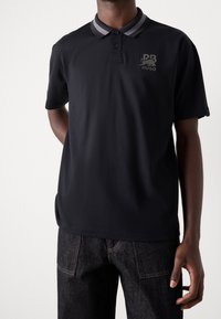 Zwarte polo shirt van katoen, met een bicolor kraag, twee knopen en een logo op de linkerborst. Draagt bij donkere jeans.
