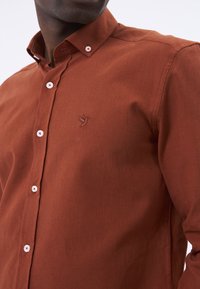 Camisa de botón en color óxido, hecha de una tela texturada, con botones blancos y un logo bordado de un pájaro en el pecho.