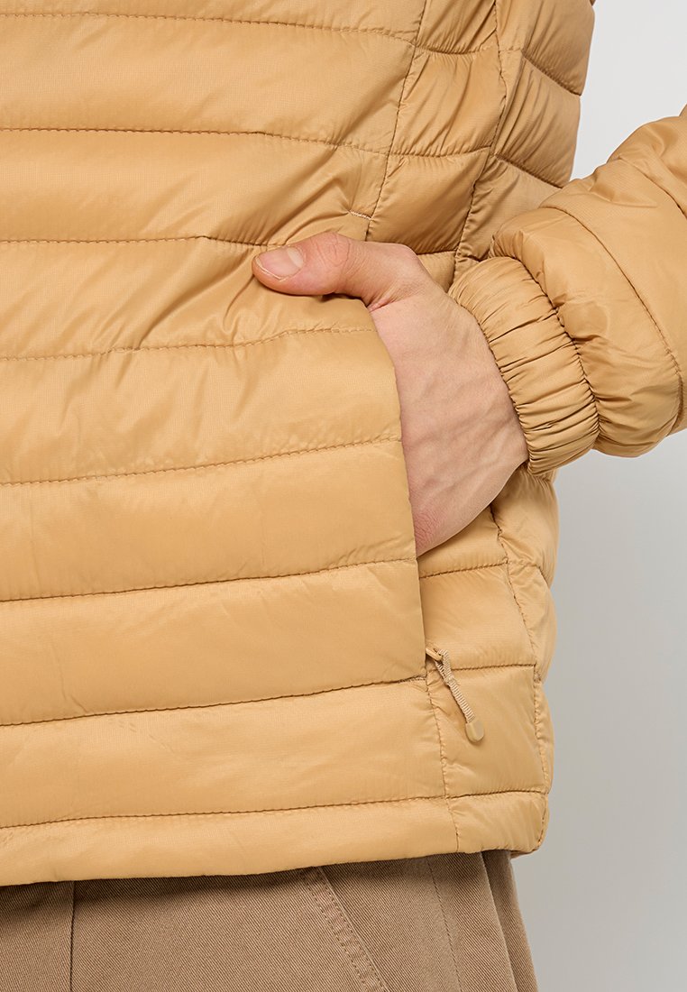 Mano in tasca di una giacca trapuntata marrone chiaro con polsini elastici e tasca laterale con zip, pantaloni beige visibili sotto.