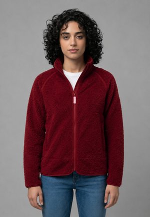 Kvinde med krøllet sort hår iført en rød fleecetrøje med lynlås og blå jeans, stående foran en neutral baggrund.