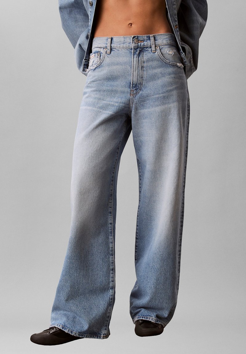 Jean délavé bleu clair, taille basse et jambes larges, porté avec des chaussures noires et une veste en jean courte laissant apparaître le ventre.