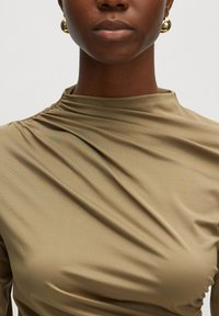 Khakifarbene Bluse mit langen Ärmeln, tailliertem Design und gerafftem Stoff am Ausschnitt, kombiniert mit goldenen Creolen.