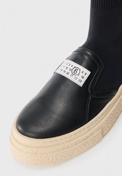 Svart lädersko med en tjock beige sula och en tygankel. Har en vit etikett med siffror, vilket ger en unik designelement.