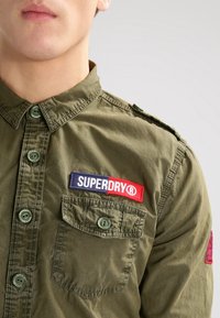 Camicia button-up verde oliva con colletto, dotata di due tasche sul petto e un'etichetta logo "SUPERDRY" blu e rossa sul lato sinistro.