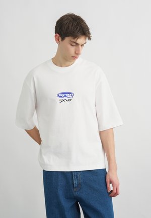 Νέος άνδρας που φοράει λευκό φαρδύ μπλουζάκι με μπλε λογότυπο και κείμενο, συνδυασμένο με φαρδιά μπλε τζιν, κοιτάζει προς τα κάτω.