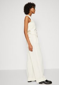 Top blanc sans manches avec un design structuré et des boutons sur le côté, associé à un pantalon crème à jambes larges et des chaussures noires brillantes.