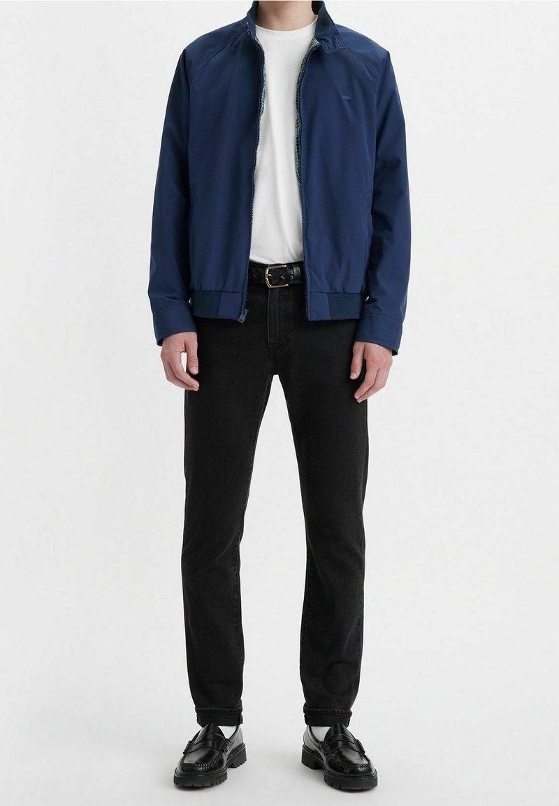 Personne debout de face, portant une veste bleue, un t-shirt blanc, un jean noir, une ceinture noire et des chaussures noires, sur un fond uni.
