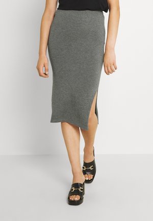 Vrouw die loopt en een grijze gebreide midi-rok met een zijsplit draagt, gecombineerd met zwarte open-toe slip-on sandalen met gouden gespen.