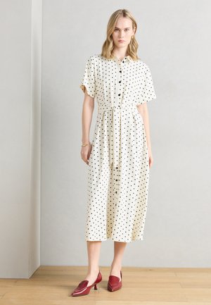 Γυναίκα που στέκεται σε εσωτερικό χώρο φορώντας άσπρο midi φόρεμα με μαύρες πουά, κοντά μανίκια, κουμπιά μπροστά, δετή μέση και κόκκινα παπούτσια με χαμηλό τακούνι.